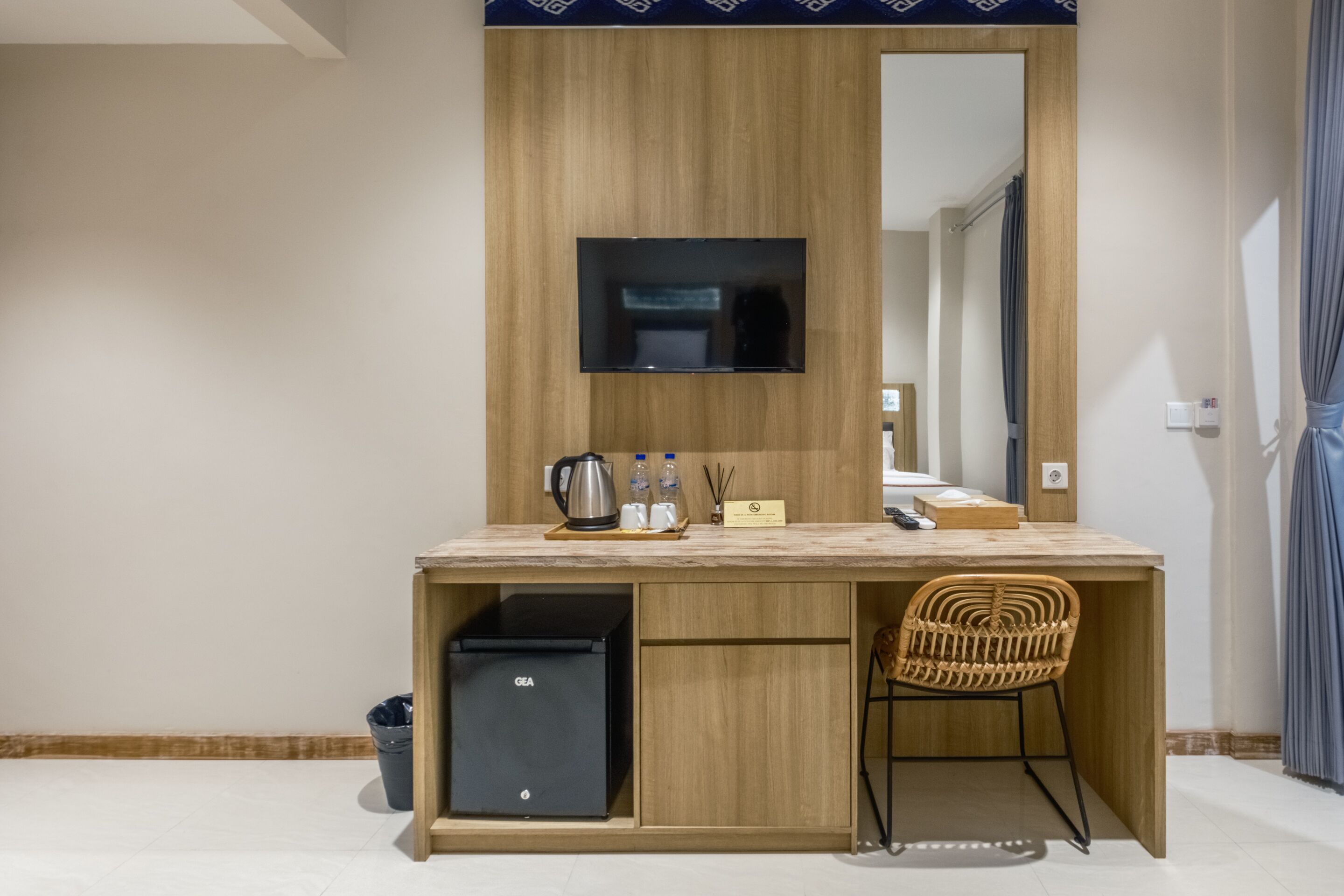 Habitación de hotel compacta con escritorio de madera, televisor de pantalla plana, refrigerador negro pequeño, silla de mimbre y un espejo grande.