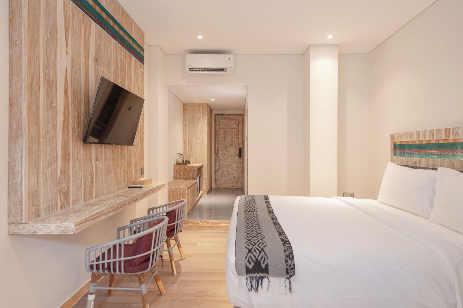 A hotel room with a bed, a TV mounted on the wall, a desk with chairs, and a wooden accent wall.