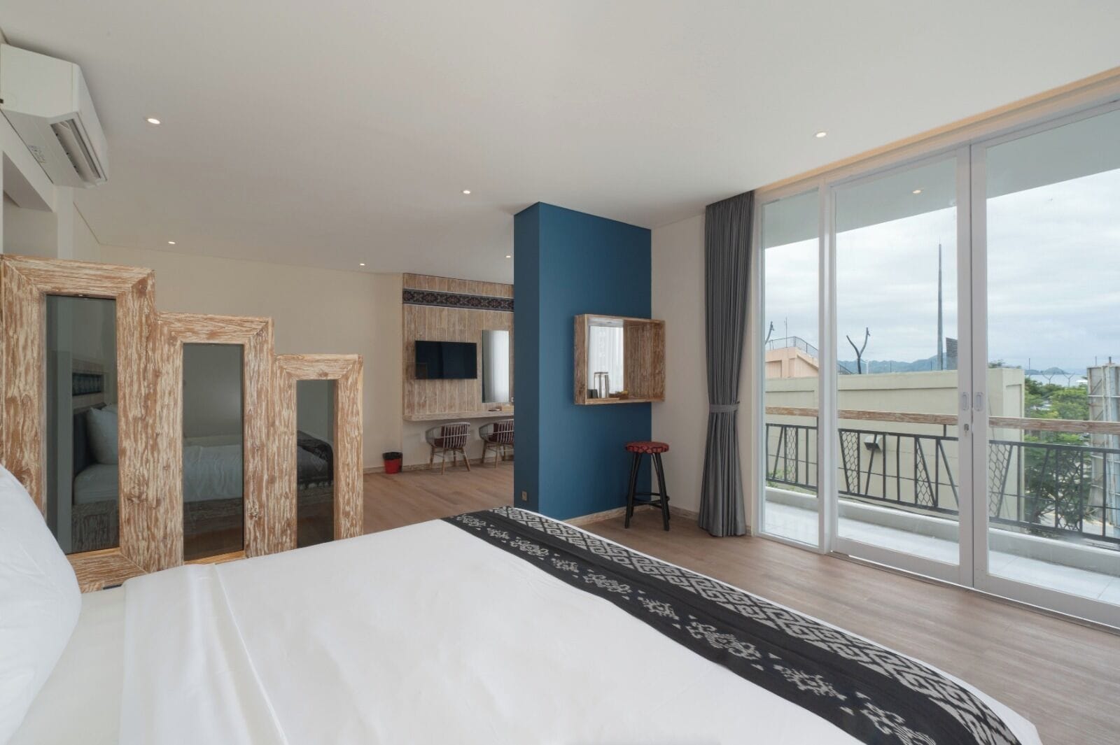 A modern hotel room with a large bed, a balcony with a view, a TV, and a mirror.