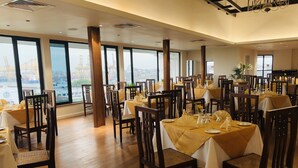 A dining area with tables set for four, large windows offering a view of a construction site, and a chandelier hanging from the ceiling. - Grand Oriental Hotel (Colombo)