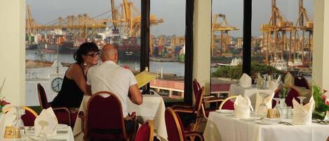 A restaurant with tables set for dining, large windows offering a view of a harbor with cranes.