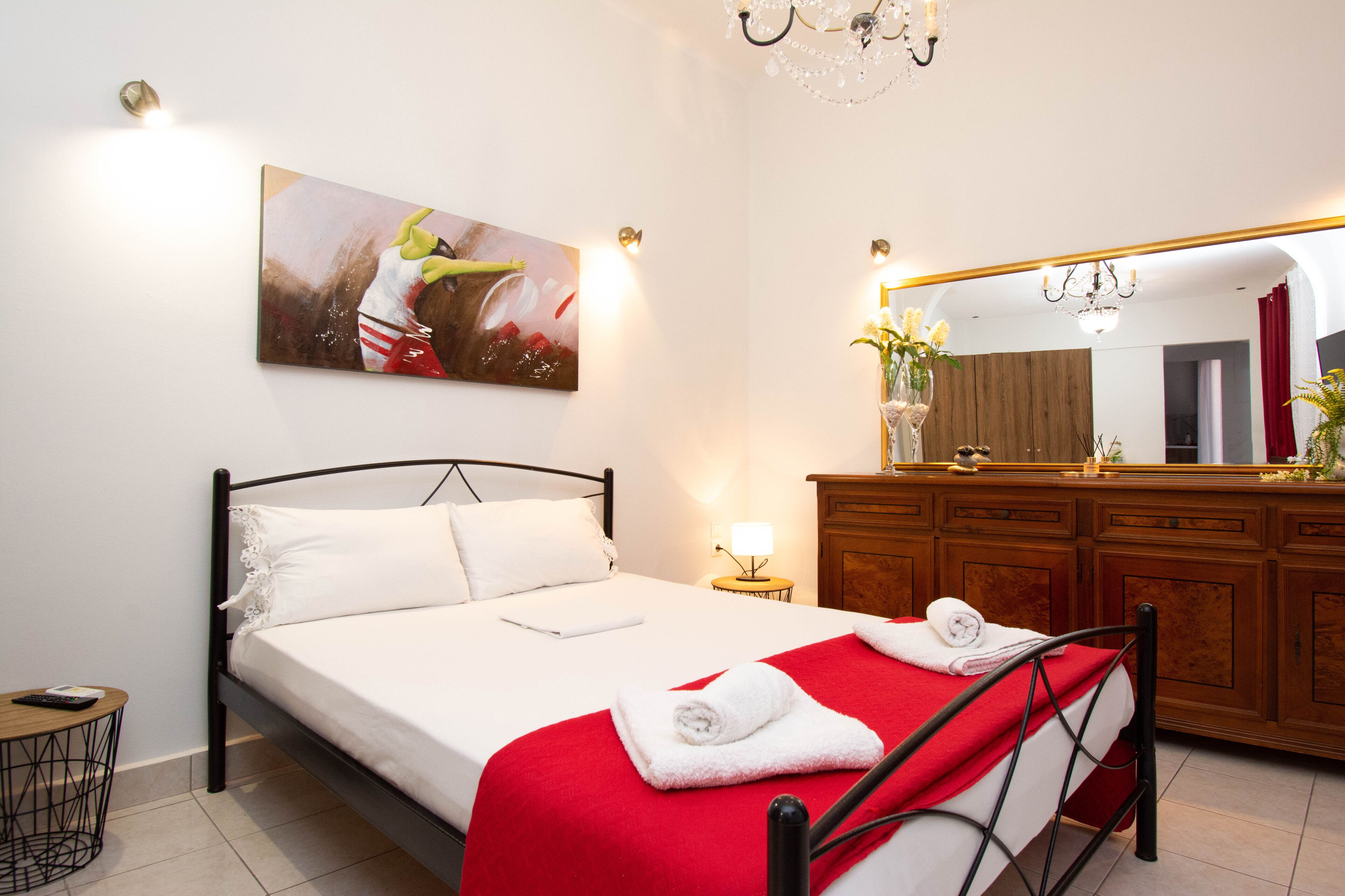 Een net opgemaakt bed met een rode deken, een nachtkastje met een lamp, een houten dressoir en een schilderij aan de muur.