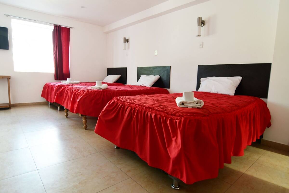 A hotel room with two beds, each with a red bedspread and white pillows, a small table, and a window with red curtains.