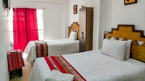 Habitación de hotel con dos camas, cada una con cabecero de madera, ropa de cama blanca y una manta roja estampada. Se observa un armario y un espejo en la pared, además de una cortina roja.