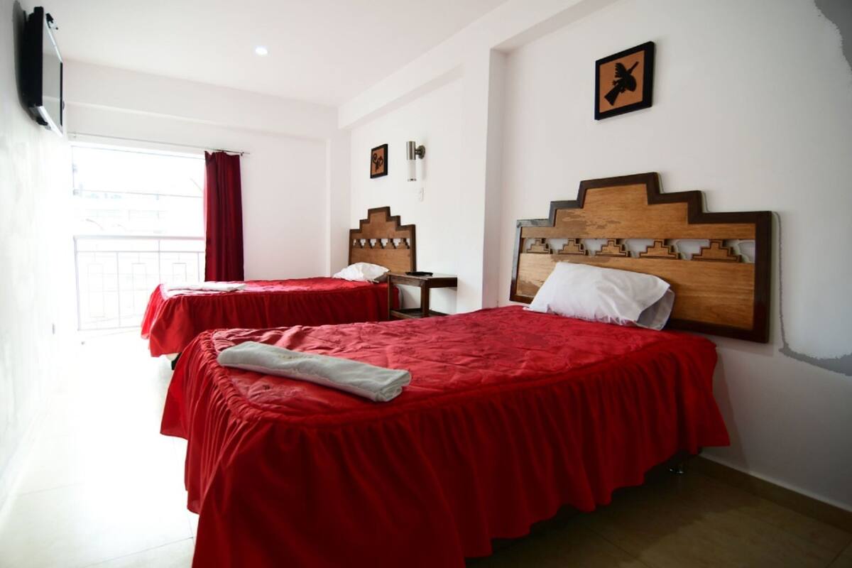 A hotel room with two beds, each with a red bedspread and white pillows. There is a wooden headboard on the right bed, and a small table with a lamp between the beds. The room has white walls and a window with red curtains.