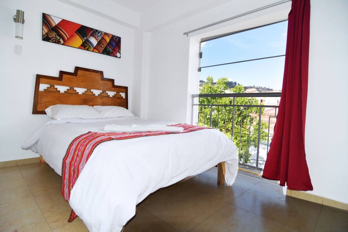 A bedroom with a wooden headboard, a large bed with white bedding, a red and white striped blanket, a window with a red curtain, and a cityscape view.