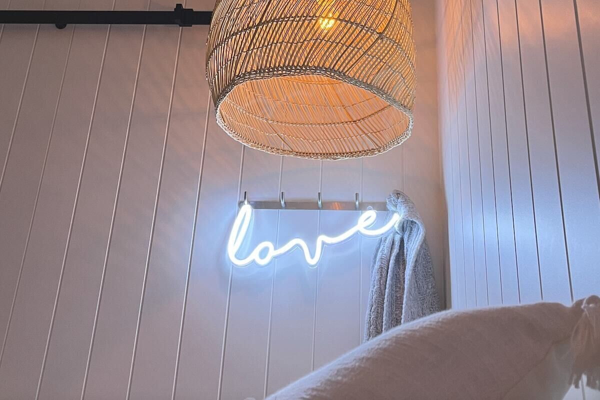 Una habitación con pared de madera blanca, lámpara de metal trenzado y un letrero de neón que dice 