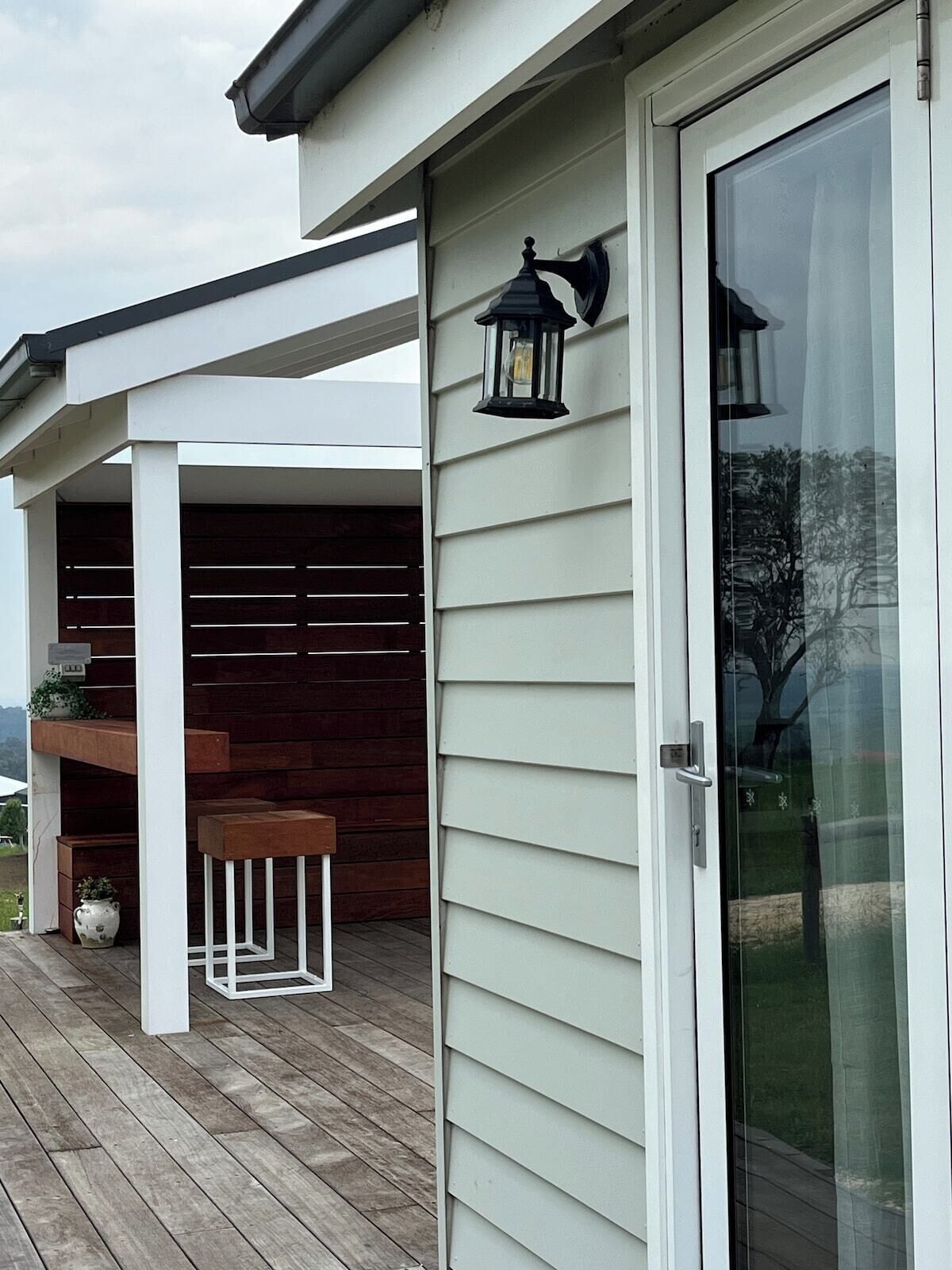 A covered outdoor area with a wooden deck, a white pergola, and a glass door with a sliding mechanism.