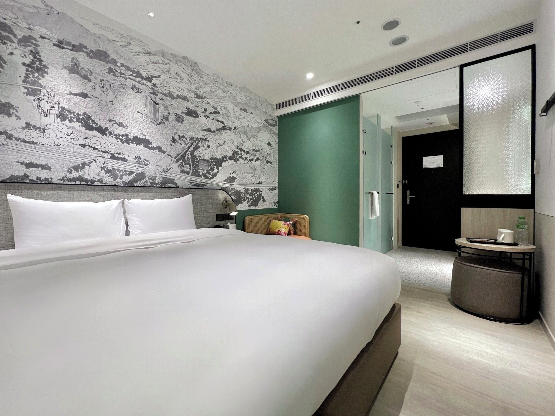 A modern hotel room with a large bed, a green accent wall, a small seating area, and a bathroom visible through a glass door.