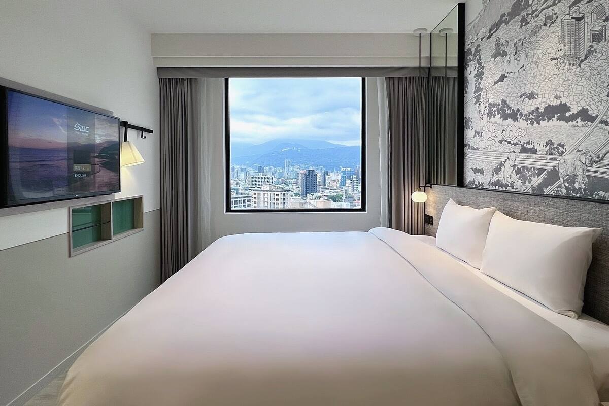 A hotel room with a large bed, a TV mounted on the wall, a city view through the window, and a decorative tapestry on the wall.