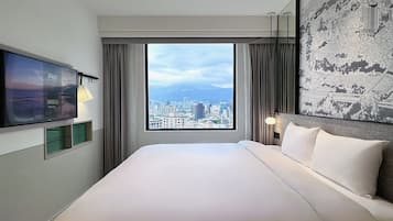 A hotel room with a large bed, a TV mounted on the wall, a city view through the window, and a decorative tapestry on the wall.