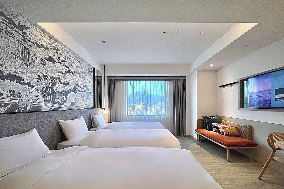 A hotel room with two beds, a sofa, a TV, and a large window with a view.