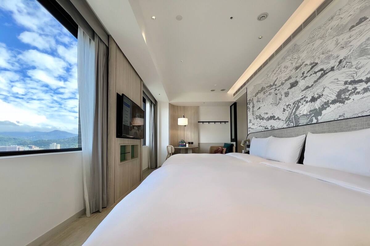 A modern hotel room with a large bed, a TV, a desk, and a view of the cityscape.