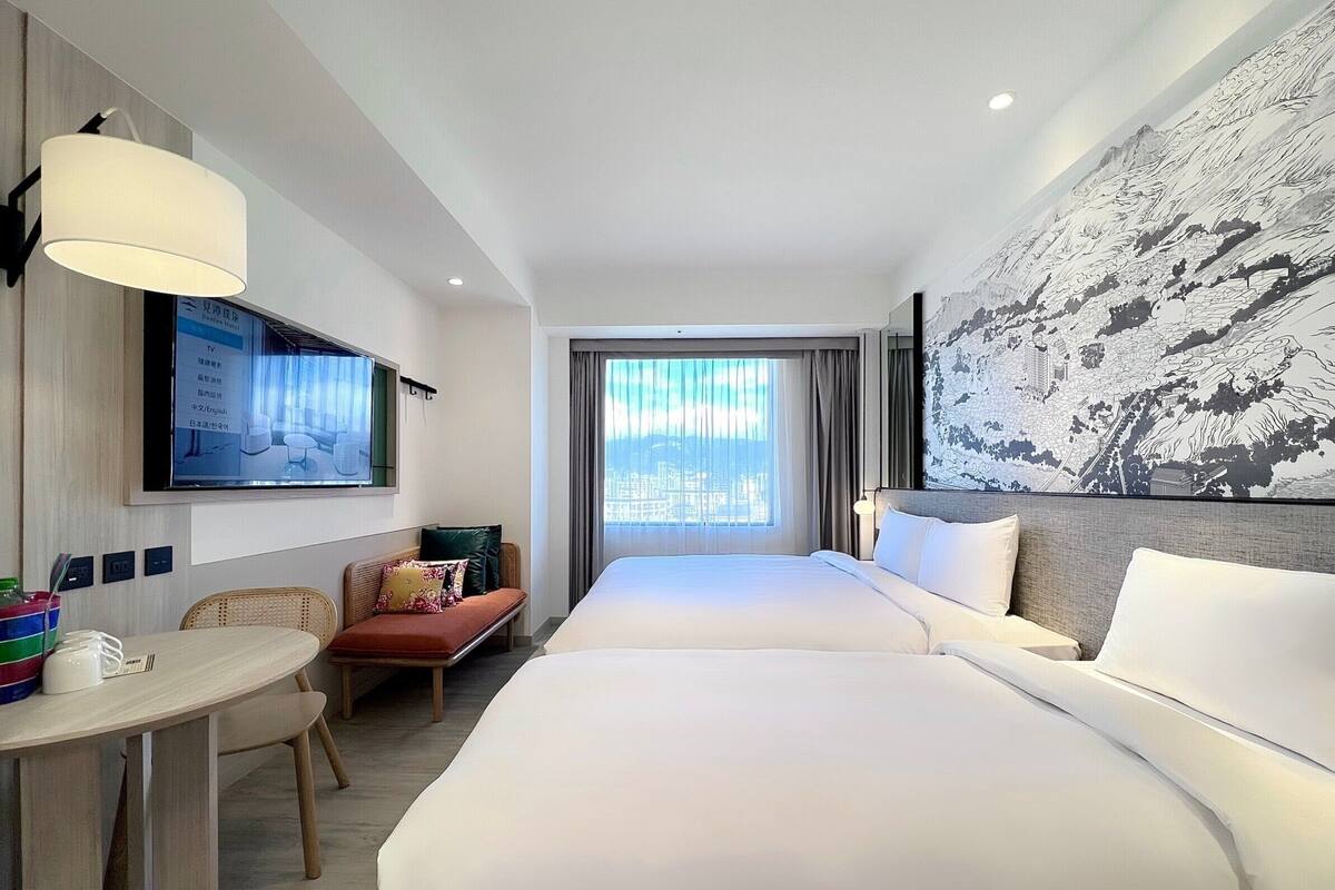 A hotel room with a large bed, a TV mounted on the wall, a desk with a chair, and a window with curtains.