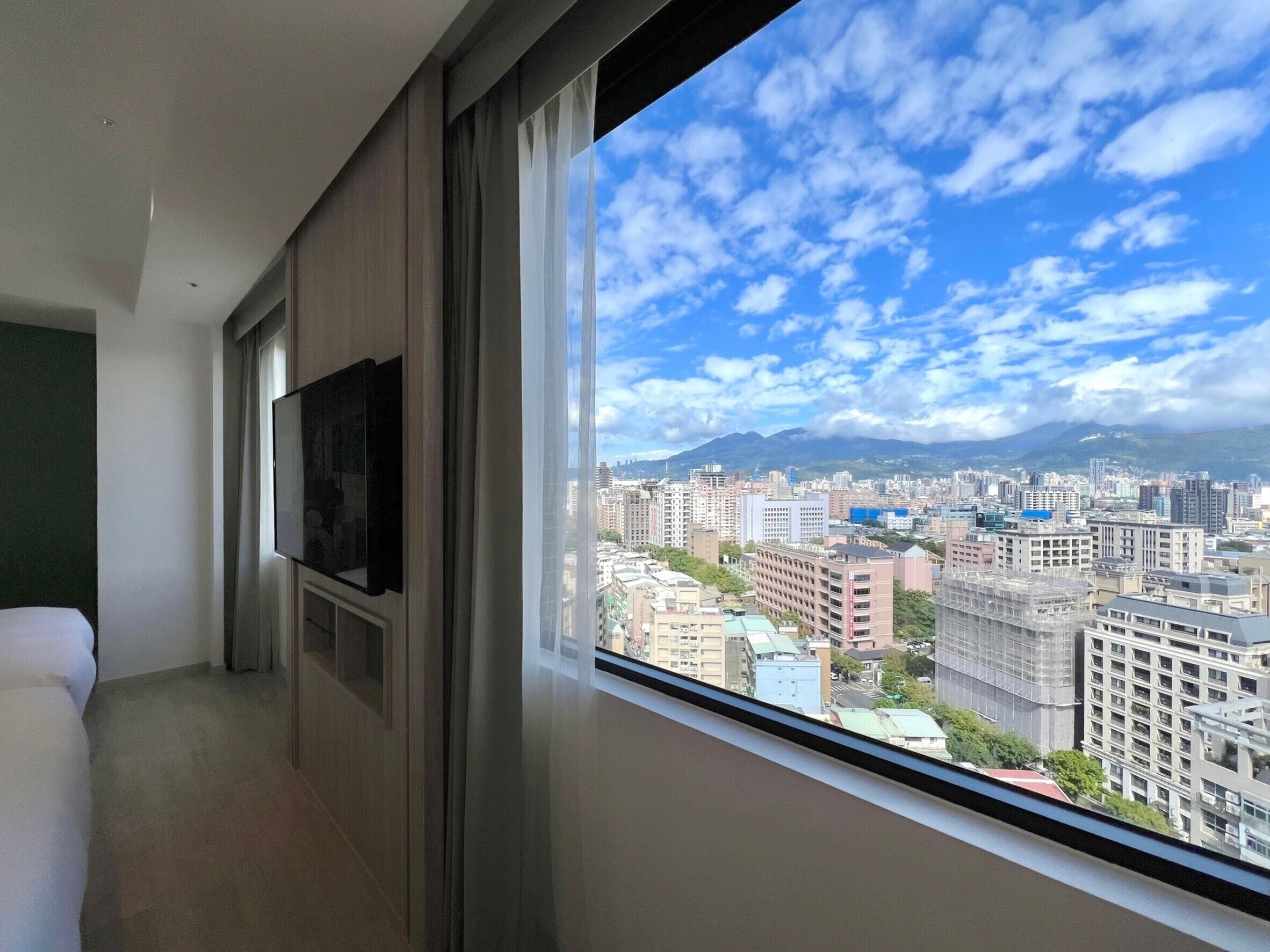 A hotel room with a large window offering a cityscape view, a flat-screen TV mounted on the wall, and a bed with a white headboard.