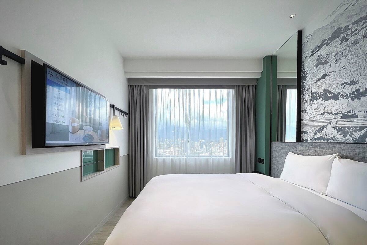 A hotel room with a large bed, a television mounted on the wall, and a city view through the window.