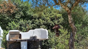 A rustic outdoor seating area with a stone table and a white wall backdrop, surrounded by dense greenery. - Casa Luisa With Pool (Alghero)