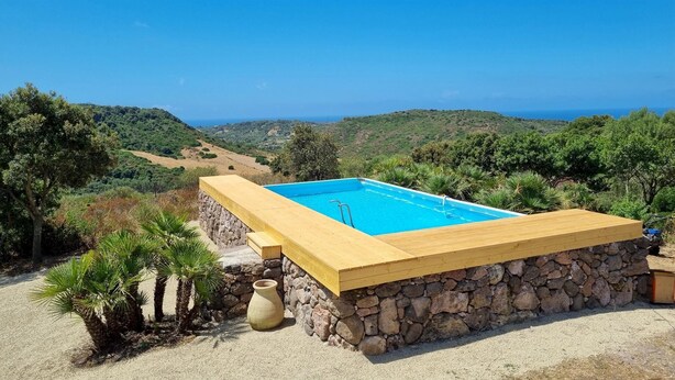 A rectangular swimming pool with a stone and wooden deck, surrounded by a gravel area and palm trees, overlooking a hilly landscape and the sea. - Casa Luisa With Pool (Alghero)