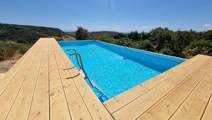 A wooden deck leading to a rectangular swimming pool with clear blue water, surrounded by greenery and hills in the background. - Casa Luisa With Pool (Alghero)