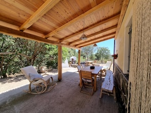 Une terrasse extérieure couverte, aménagée avec du mobilier en bois, notamment une table et des chaises, ainsi qu’un fauteuil à bascule en osier.