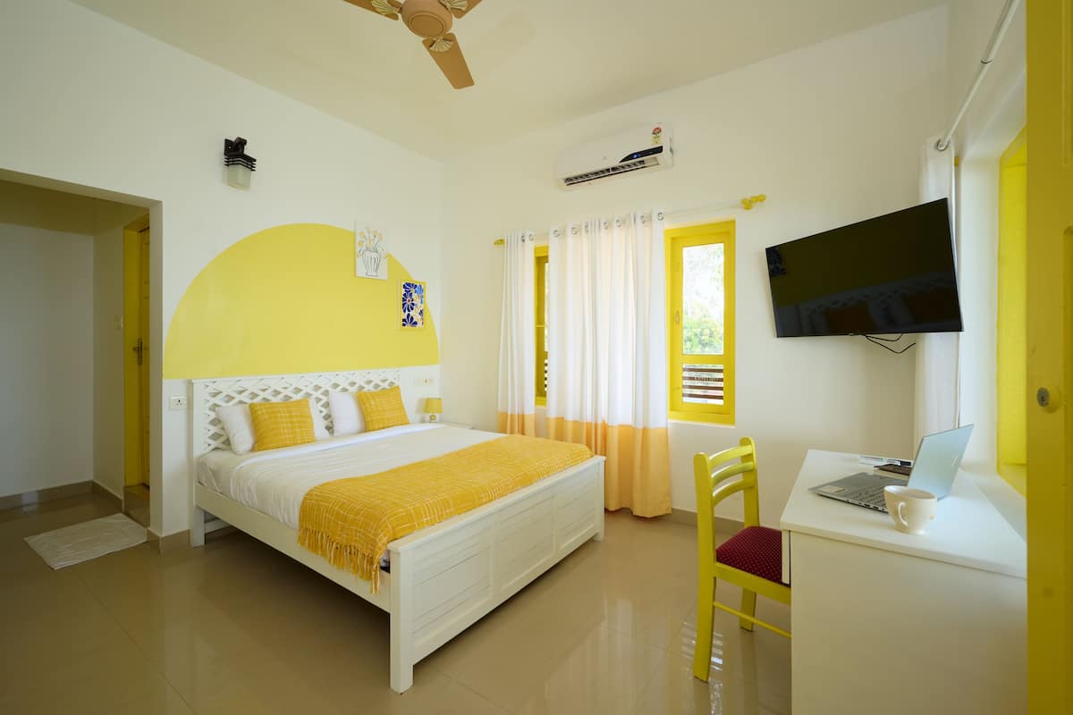 A bedroom with a bed, a desk with a laptop, a TV, and a window with curtains.