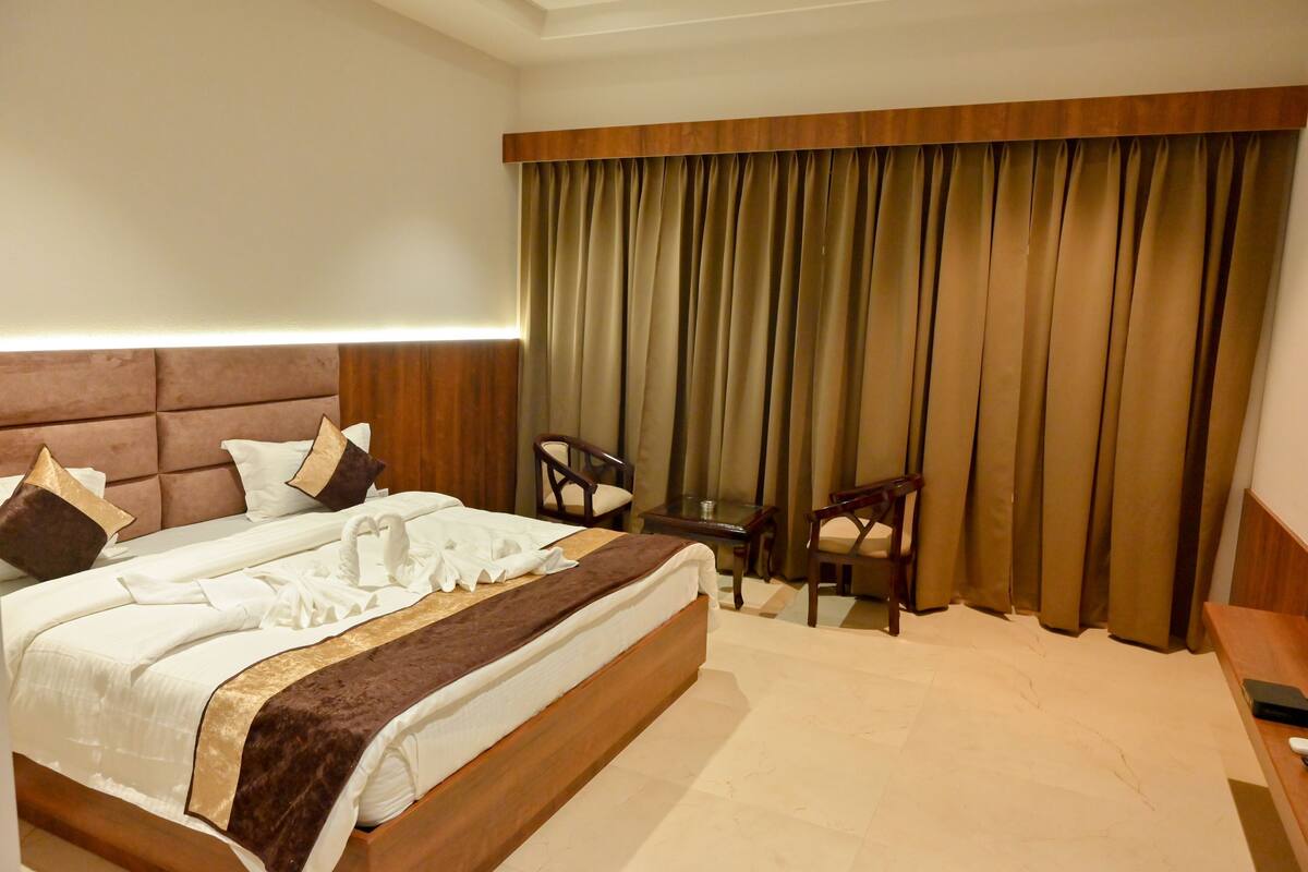 A hotel room with a large bed, a wooden headboard, a desk with a chair, and a large window with curtains.