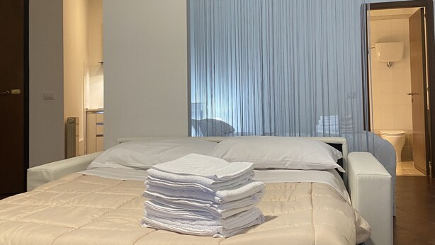 A neatly made bed with a stack of white towels, a bedside chair, and a bathroom visible through a window with sheer curtains. - House Valentina (Perugia)