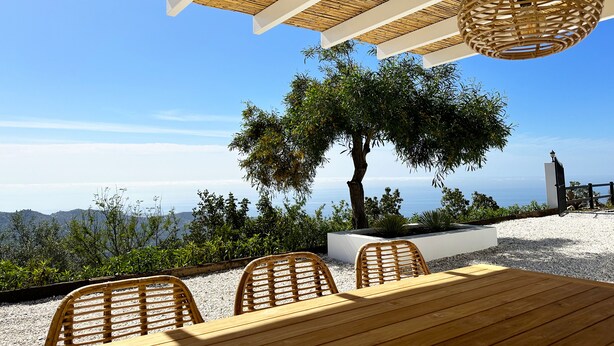 Un espacio al aire libre con mesa y sillas, techo de paja y vista al mar.