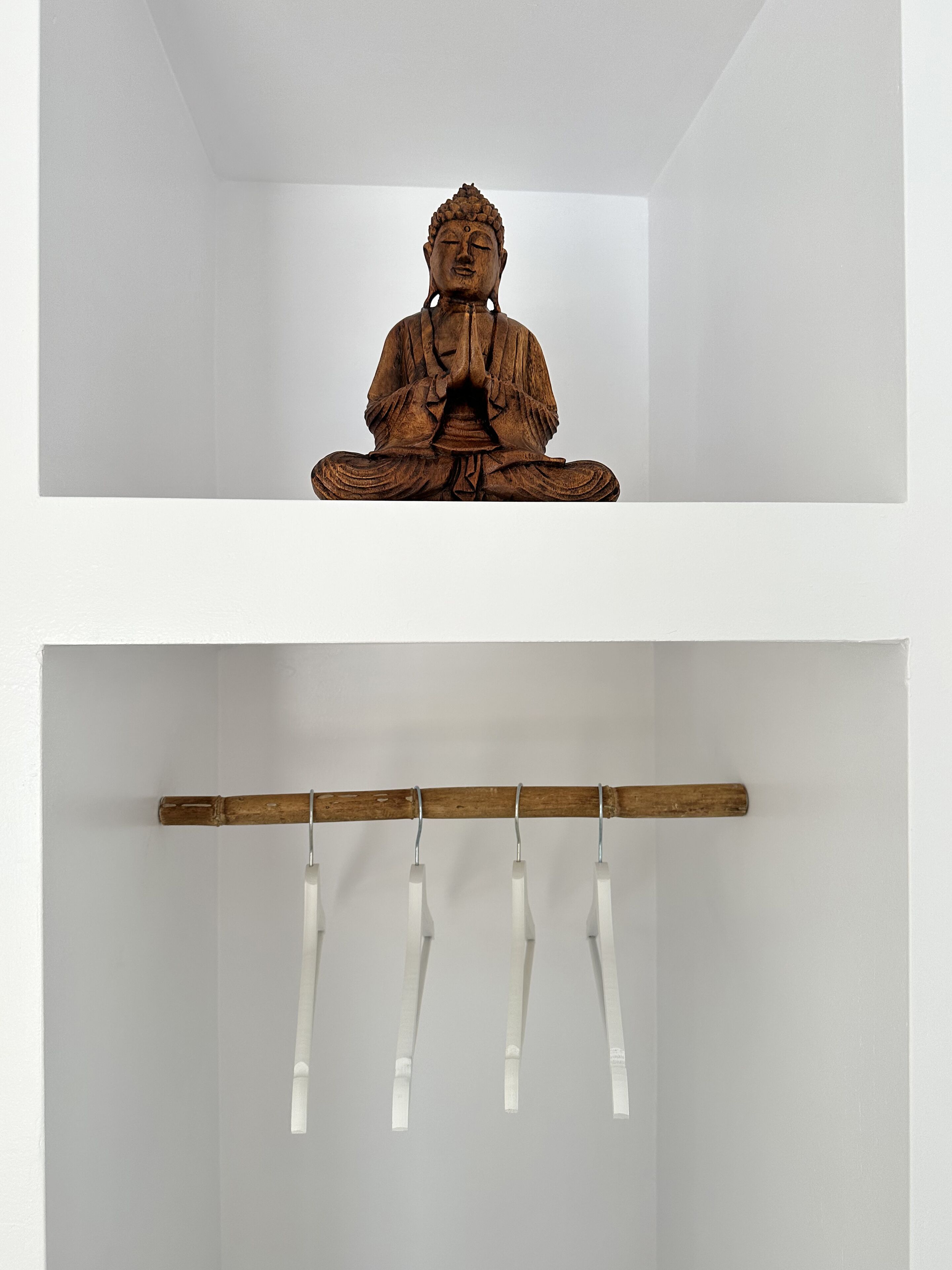Une statue en bois représentant une figure assise, dans une pose méditative, est posée sur une étagère. En dessous, une tige en bois, à laquelle sont attachées plusieurs lanières blanches, est suspendue au plafond.
