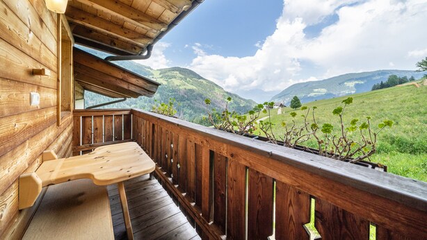 Un balcon en bois avec une table en bois, une balustrade et une vue sur des collines verdoyantes et des montagnes.