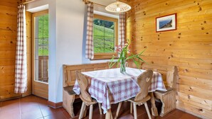 L’intérieur d’un chalet en bois comprend une table à manger, des chaises, une fenêtre avec des rideaux, un luminaire suspendu et un tableau encadré accroché au mur.