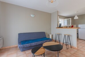 Un espace de vie compact comprenant un canapé, deux tabourets, une petite table, un coin cuisine et un luminaire fixé au plafond.