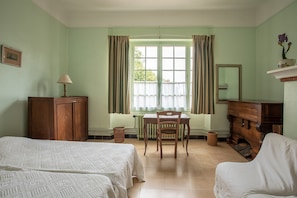 Ein Zimmer mit zwei Betten, einem Holztisch mit Stuhl, einem Schrank, einer Heizung und einem Fenster mit Vorhängen.