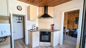 Eine kleine Küche mit Herd, Backofen und Kaffeemaschine. An der Wand hängt eine Uhr, und die Decke besteht aus Holz.