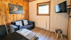 Ein gemütliches Zimmer mit Holzbalkendecke, einer Couch mit gemusterten Kissen, einem an der Wand montierten Fernseher und einem Fenster mit Blick auf Grünflächen.