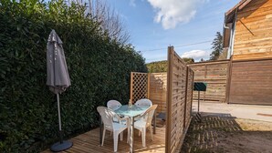 Eine Holzterrasse mit einem Glastisch und Stühlen, umgeben von einem Holzzaun und einer Hecke.