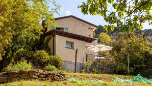 A two-story house with a stone wall in the foreground, a pergola with a beige umbrella, and a garden with various plants and trees. - Holiday Home 'Il Pescino' with Private Terrace and Wi-Fi (Pietrasanta)