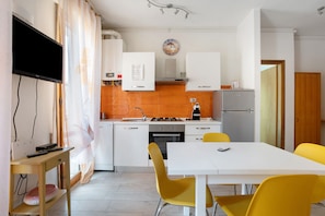 Une cuisine moderne avec des meubles blancs, une cuisinière et un réfrigérateur. On y trouve une table à manger avec des chaises jaunes et un téléviseur fixé au mur.