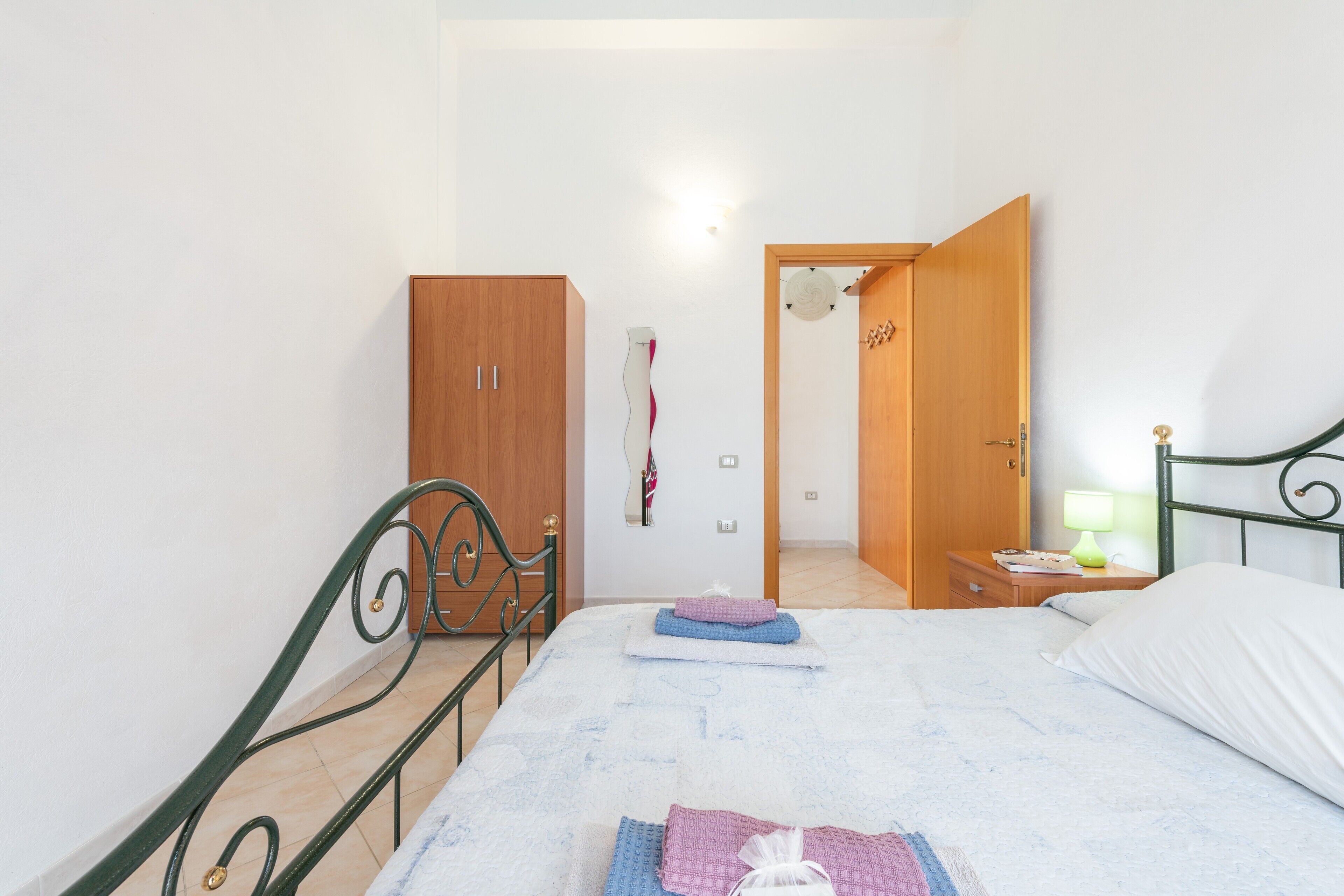 A bedroom with a bed, a wardrobe, a nightstand with a lamp, and a door leading to another room.