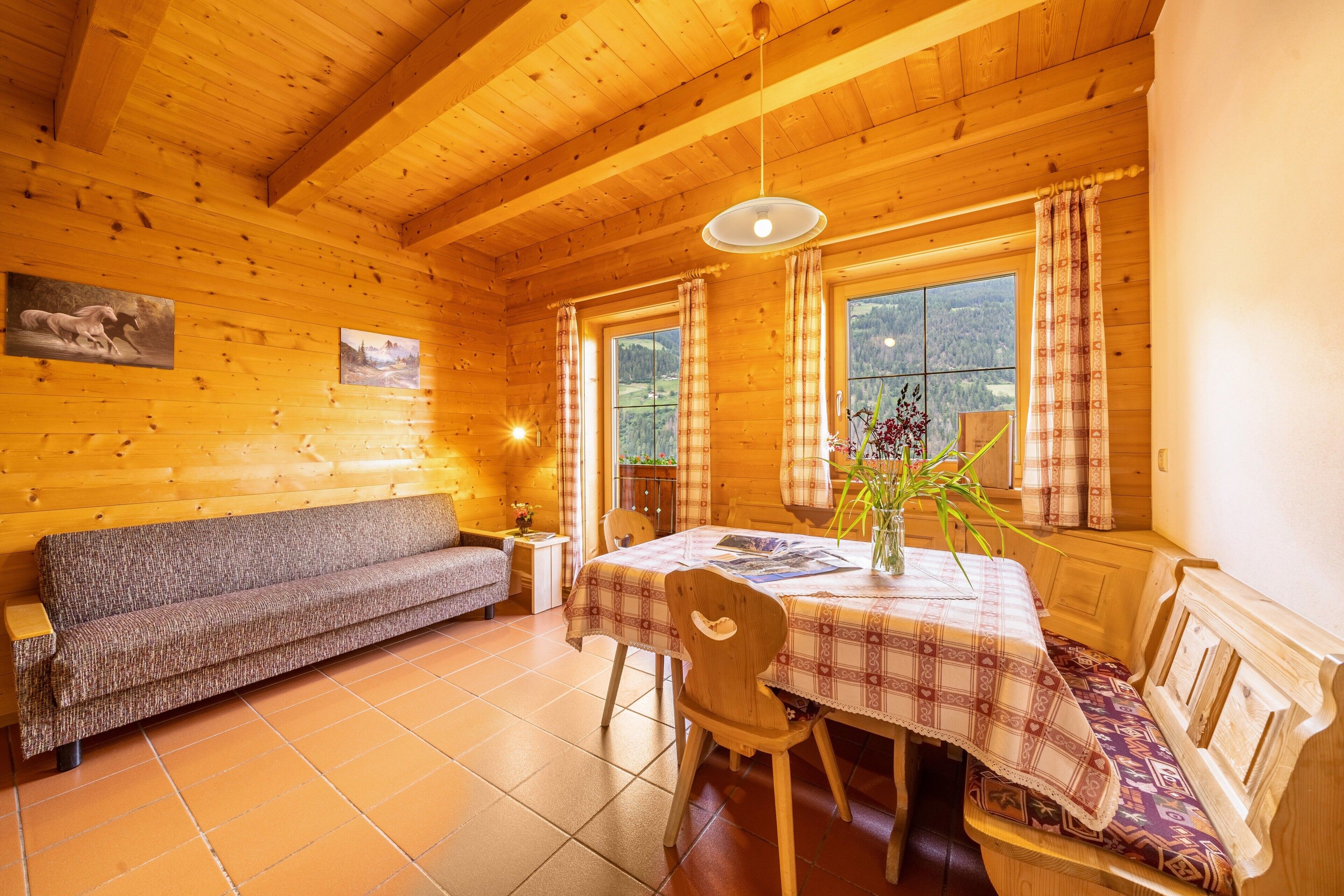 A wooden cabin interior with a sofa, dining table, chairs, and a view through the windows.