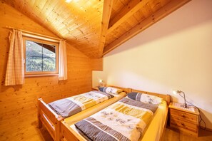 Une chambre dans une cabane en bois, avec deux lits, une fenêtre avec des rideaux, une table de chevet avec une lampe, et une vue sur l’extérieur.