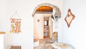 Una habitación con un arco de piedra, un adorno colgante, una silla de mimbre, una mesa redonda y un espejo.
