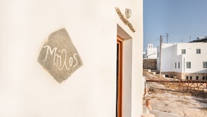 Un edificio con un letrero que dice 'Milos' en una pared blanca, una puerta de madera y una vista de otro edificio blanco al fondo.