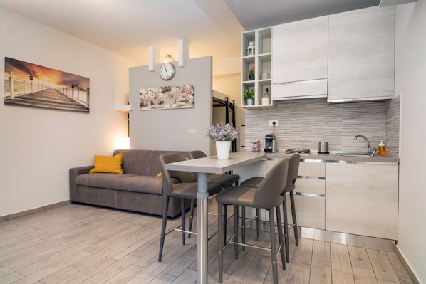A modern kitchen with a built-in island, a dining area with a table and chairs, a sofa with a cushion, and a clock on the wall.