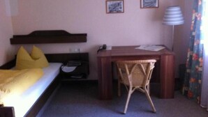 Quarto de hotel com uma cama, uma escrivaninha, uma cadeira, um abajur e duas molduras na parede.