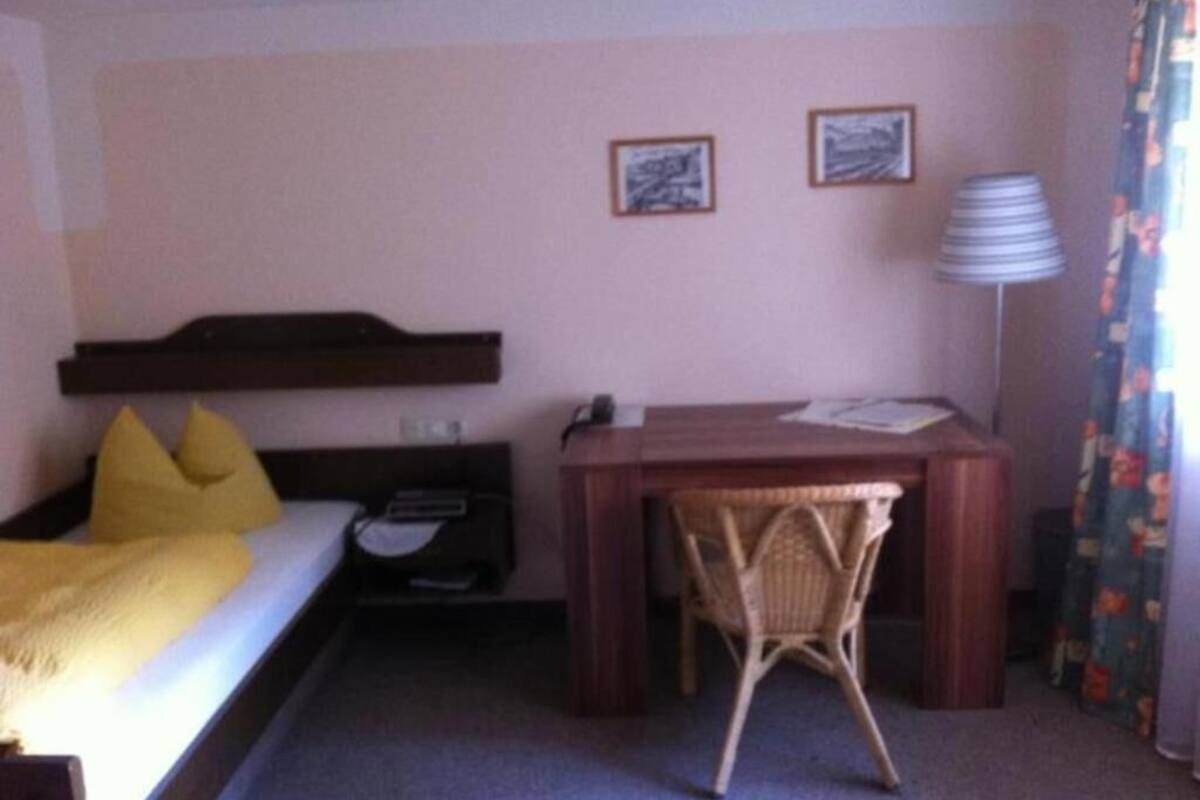 A hotel room with a bed, a desk, a chair, a lamp, and two framed pictures on the wall.