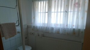 Casa de banho com sanita, cabine de duche em vidro, radiador e janela com cortinas florais.