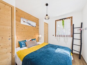 A bedroom with a wooden headboard, a bed with a blue and yellow blanket, a painting above the bed, a hanging light fixture, a window with sheer curtains, and a ladder against the wall.