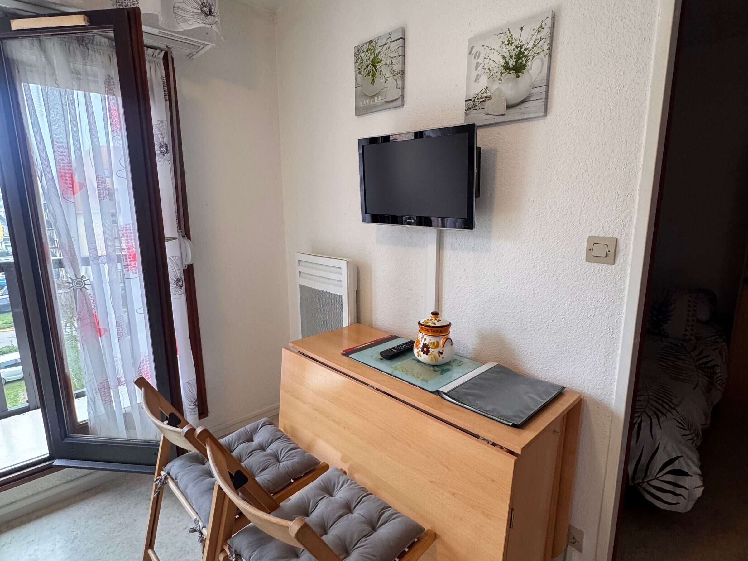 A room with a wooden desk, a television mounted on the wall, a window with curtains, and a bed with a patterned bedspread.
