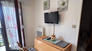 A room with a wooden desk, a television mounted on the wall, a window with curtains, and a bed with a patterned bedspread.